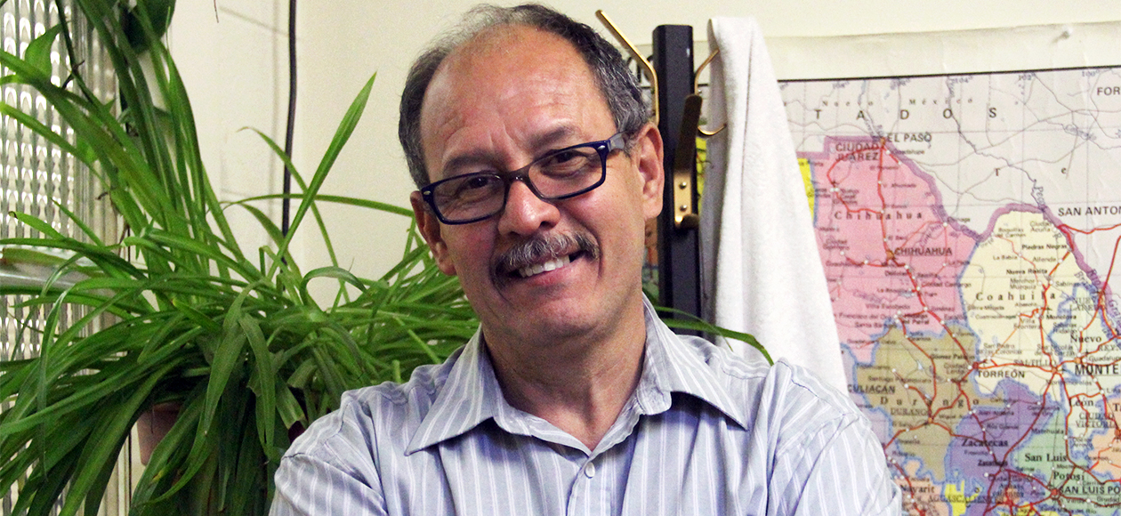 Dr. Sergio Inestrosa, with his dark-rimmed glasses and gray mustache, smiles for a photo in his office. He is wearing a blue collared shirt and is standing in front of a map of the world with a bright, green plant perched on a bookshelf behind him.