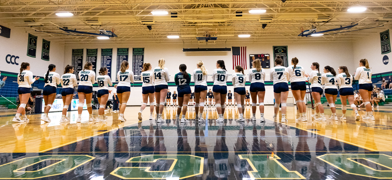 The women's volleyball team stands facing away from the camera, on the end line, for their pre-match lineup introductions.