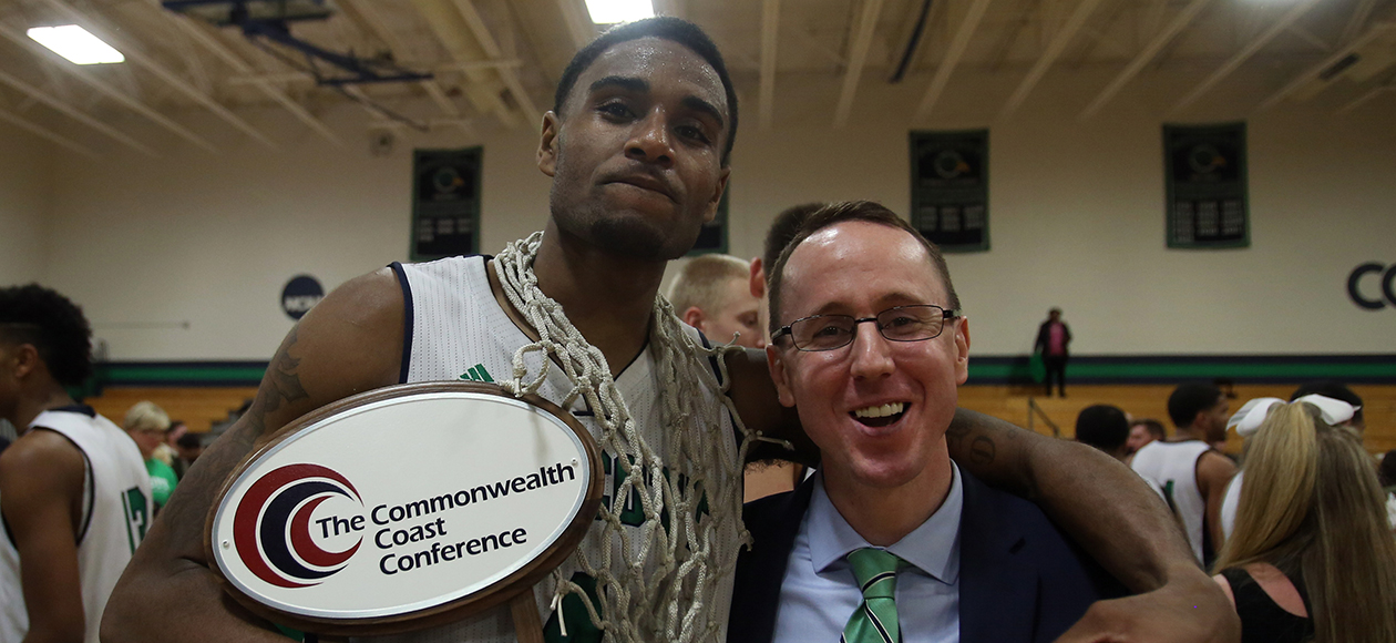 Daquan Sampson, with a basketball net draped around his neck, holds the Commonwealth Coast Conference trophy in his right arm while putting his left arm around men's basketball assistant coach Luke Richards as they smile for a photo to celebrating winning the conference championship in the year 2016.