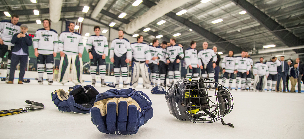 Hockey gloves and a stick lay in the foreground while the men's ice hockey team stands in the back - out of focus - in a line awaiting the CCC championship presentation at Raymond J. Bourque Arena.