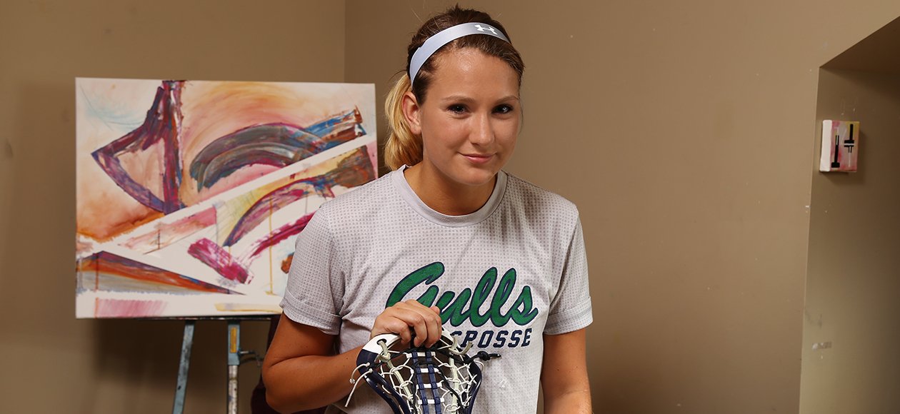 Senior women's lacrosse player Kendra Walkers poses with a smile in the art studio next to one of her paintings while holding her lacrosse stick.